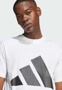 Witte katoenen t-shirt met een groot zwart trefoil-logo op de voorkant. Korte mouwen en een ronde hals. Vriendelijke textuur.