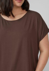 Vrouw met schouderlang donker haar, gekleed in een losvallende, korte mouw bruine top tegen een effen lichte achtergrond.