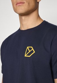 Mornarji modra bombažna majica z rumenim geometrijskim logotipom na levem prsnem predelu. Majica ima okroglo ovratnico in kratke rokave.