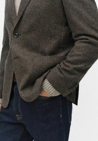 Brun blazer med fiskbensmönster och skräddarsydd passform, försedd med svarta knappar och en subtil lyster, över en ljusgrå tröja och mörka jeans.