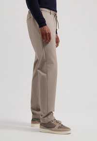Pantaloni bej cu talie elastică și șnur, având buzunare laterale și un design slim fit, asortați cu adidași gri.