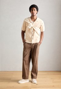 Kortærmet beige skjorte med knapper og tekstureret stof, parret med brune vidde bukser og hvide sneakers. Accenttrøje hængt over skuldrene.