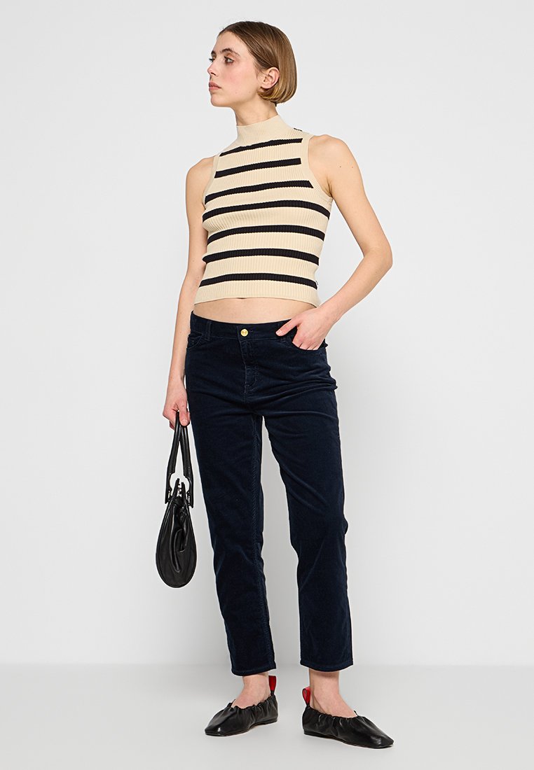Frau trägt ein ärmelloses Oberteil mit beigen und schwarzen Streifen, schwarze Hose, schwarze Ballerinas, hält eine schwarze Handtasche und steht vor einem weißen Hintergrund.
