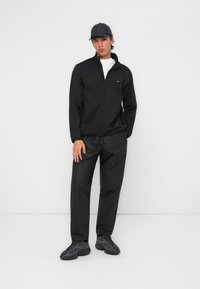 Black zip-up jacket, white t-shirt, black trousers, and dark sneakers; model stands against a white background, featuring a simple, clean design.