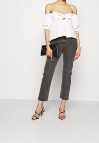 Donna che indossa un top bianco scollato, jeans grigi scuri a vita alta, tacchi neri con cinturino, mentre tiene una pochette nera con catena dorata su uno sfondo bianco.
