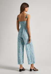 Lichtblauwe jumpsuit met bloemenpatroon, gesmokt lijfje en wijde broekspijpen. Voorzien van verstelbare schouderbanden en franje-accenten. Zwarte blokhak sandalen.