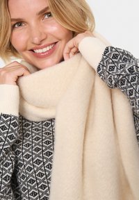 Krém színű sál, puha, texturált anyagból készült, a nyak köré terítve, mintázott fekete-fehér pulóverrel párosítva.