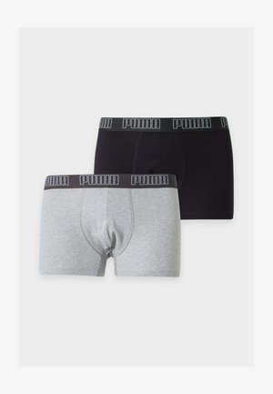 Homme mince à la peau foncée debout pieds nus portant un boxer gris Puma, une main dans le dos, sur un fond clair uni.