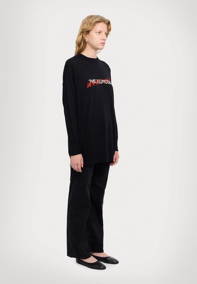 Chemise noire à manches longues avec un motif textuel graphique rouge et blanc, associée à un pantalon large noir et des chaussures plates noires. Design simple, style décontracté.