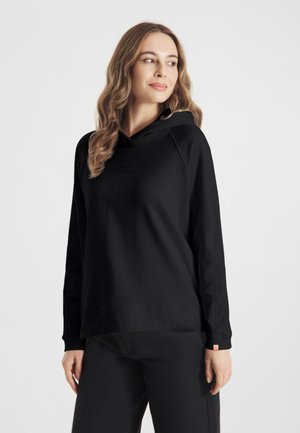 Vrouw met golvend haar, gekleed in een zwarte hoodie en zwarte broek, staand en iets zijwaarts kijkend tegen een effen lichte achtergrond.