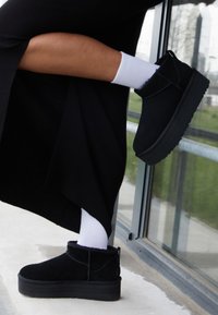 Botines de ante negros con suela gruesa, que cuentan con una tira en la parte trasera, usados con calcetines blancos hasta el tobillo. Visibles sobre una superficie lisa cerca de una ventana.