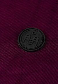 Głęboki burgundowy materiał z teksturowanym wzorem sztruksu. Okrągłe, czarne gumowe logo znajduje się wyraźnie na środku, z abstrakcyjnymi falistymi wzorami.