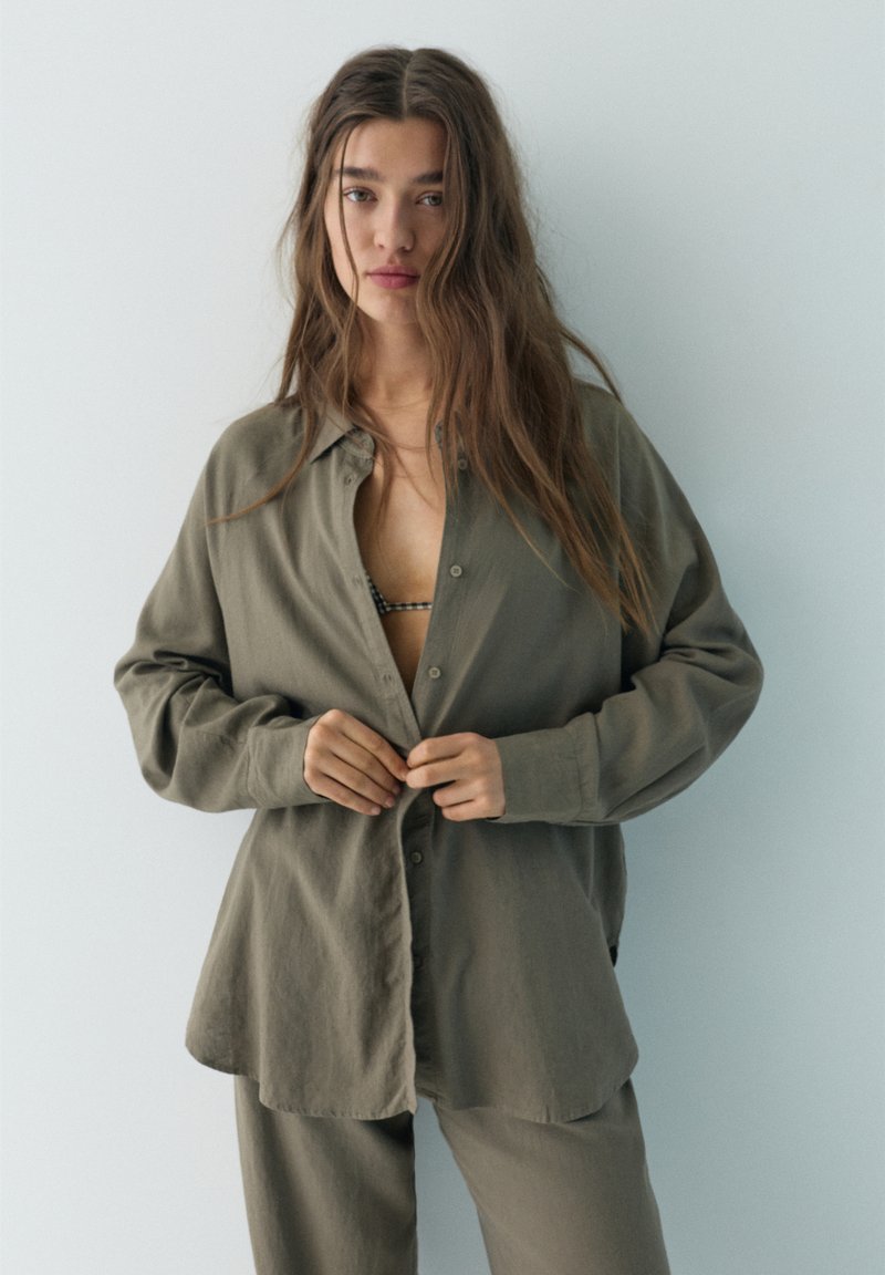 Jeune femme aux longs cheveux boutonnant une chemise surdimensionnée vert olive, portant un pantalon assorti et un soutien-gorge à carreaux noir et blanc visible.