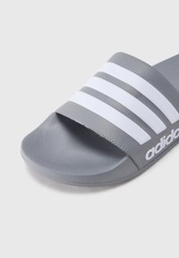 Γκρι σαγιονάρα με τρεις λευκές ρίγες στον ιμάντα, με υφή υλικού και εργονομική σόλα για άνεση. Εμφανίζεται το λογότυπο της μάρκας.