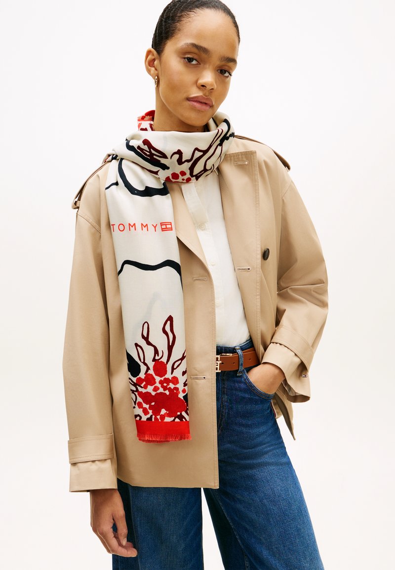 Λευκό κασκόλ με κόκκινο και μαύρο αφηρημένο σχέδιο, με το λογότυπο "TOMMY", φορεμένο πάνω από μπεζ σακάκι, συνδυασμένο με σκουρόχρωμα τζιν.