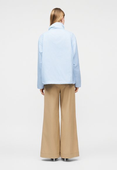 Γυναίκα που φοράει ανοιχτό μπλε σακάκι και μπεζ παντελόνι φαρδύ, στέκεται με την πλάτη σε απλό φόντο.