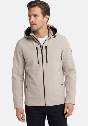 Homme portant une veste à capuche beige avec une fermeture éclair noire et des poches, assortie à un pantalon noir, regardant légèrement vers la gauche.