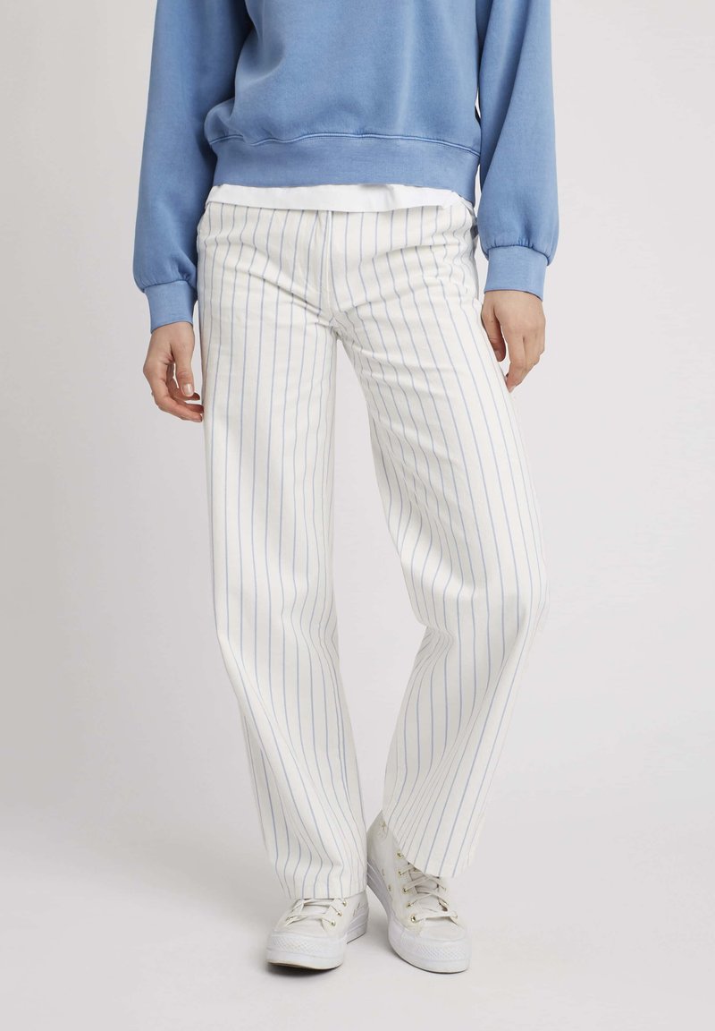 Pantalons blancs rayés avec des lignes bleues, présentant une coupe droite. Associés à des baskets blanches et un sweat-shirt bleu.