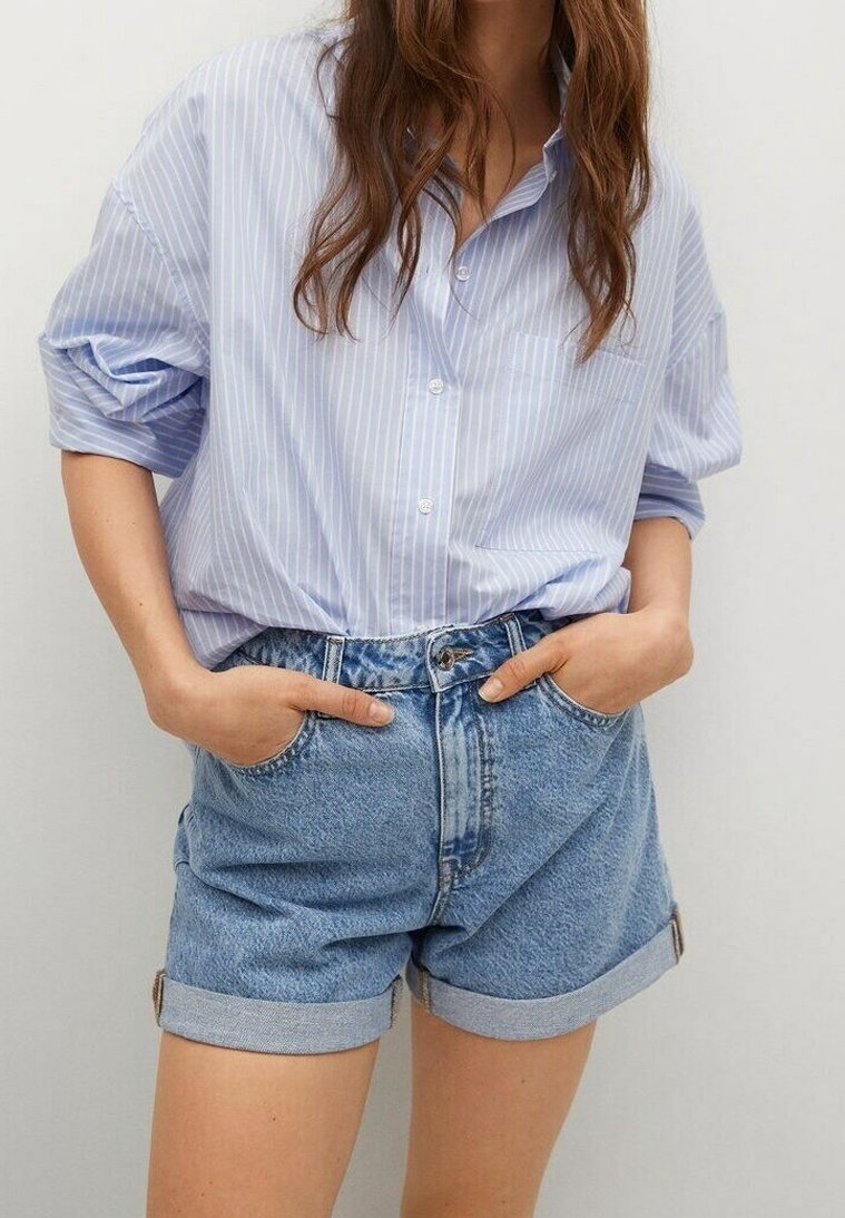 Mujer vestida con una camisa de rayas azul claro, suelta y con las mangas arremangadas, metida dentro de unos pantalones cortos de mezclilla de tiro alto y con dobladillo, con las manos en los bolsillos.