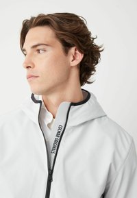 Ljusgrå huvtröja med dragkedja, med en svart accent vid halsen och logodetaljer. Slät textur och minimalistisk design.