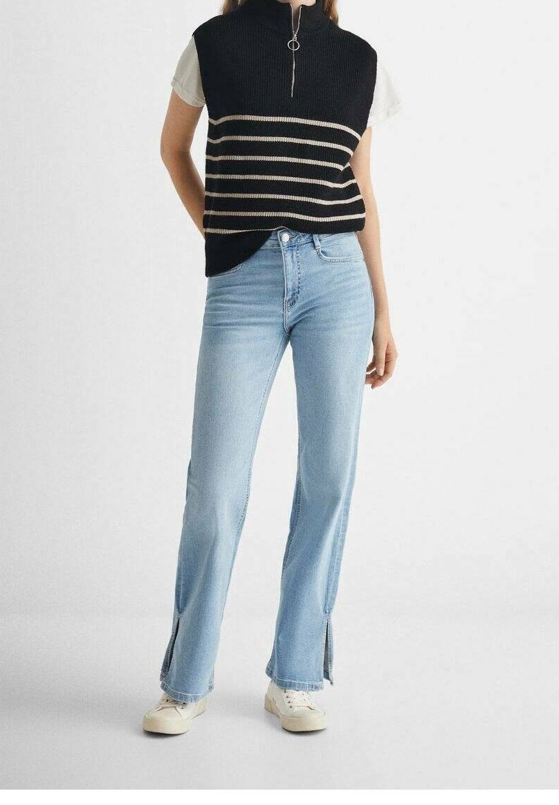 Femme portant un jean évasé bleu clair avec des fentes à l'avant, un pull sans manches rayé noir par-dessus une chemise blanche à manches courtes, et des baskets blanches.
