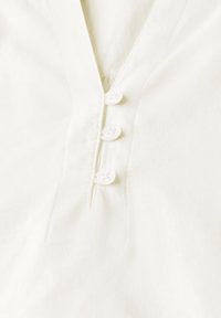 Witte katoenen blouse met een V-hals, vier witte knopen en gedetailleerde stiksels langs de knopenlijst. Gladde textuur.