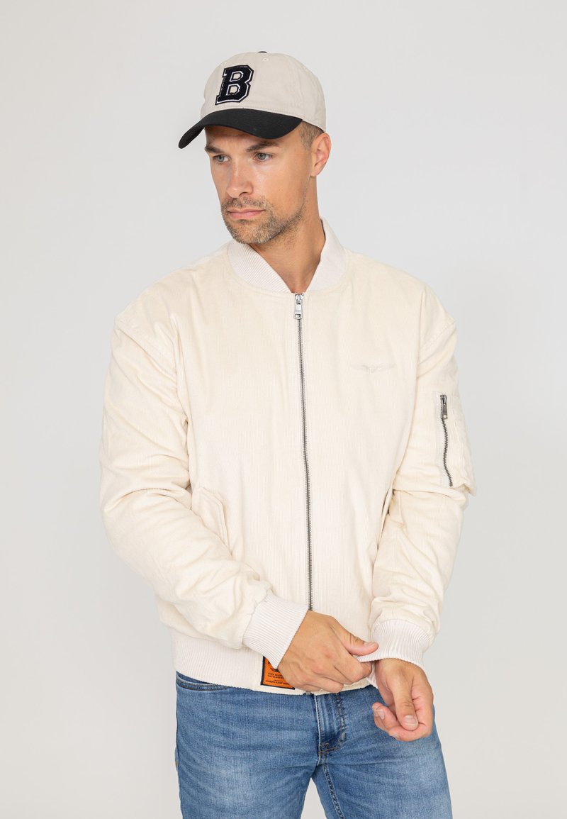 Veste bomber beige clair avec des poignets côtelés, fermeture éclair à l'avant et poches latérales. Porte avec une casquette beige comportant une lettre 'B' noire sur le devant.