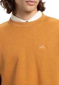 Sennepsfarvet sweatshirt lavet af blødt stof med rund halsudskæring og et lille broderet logo på venstre bryst.