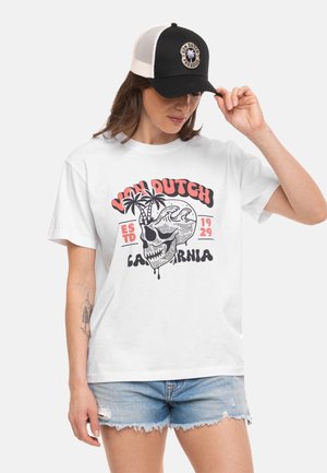 Femme portant un t-shirt blanc Van Dutch California avec un motif graphique, un short en jean bleu clair et une casquette Van Dutch noir et blanc, ajustant sa casquette avec la main droite.