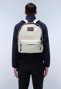 Bílý látkový batoh s černými akcenty, má nášivku norské vlajky, text "NAPAPIJRI geographic" a přední zipovou kapsu.
