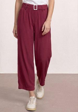 Femme portant un pantalon bordeaux taille haute à jambes larges avec ceinture et baskets blanches, marchant sur un sol beige avec un mur blanc texturé en arrière-plan.