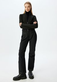 Chaqueta negra con cremallera y cuello alto, combinada con pantalones negros que presentan dos bolsillos frontales y detalles de costura sutiles. Botines negros.