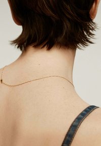 Delicada cadena de oro reposando sobre piel desnuda, con un diseño simple de eslabones y un pequeño cierre. Cabello corto y oscuro visible.