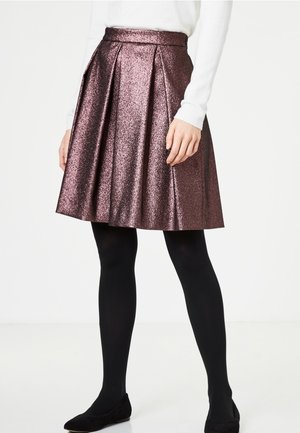 Mujer vestida con una falda plisada metálica morada hasta la rodilla, medias negras, zapatos planos negros y una camiseta blanca de manga larga, de pie sobre un fondo blanco.