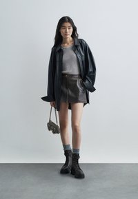 Jeune femme portant une veste en cuir noir, un haut en maille gris, une mini-jupe en cuir noir, des chaussettes grises, des bottes lacées noires et tenant un petit sac à main.