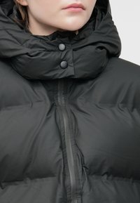 Prim-plan al unei persoane îmbrăcate cu o jachetă puffer neagră, cu guler înalt, fermoar și două nasturi negri cu capscă pe guler.