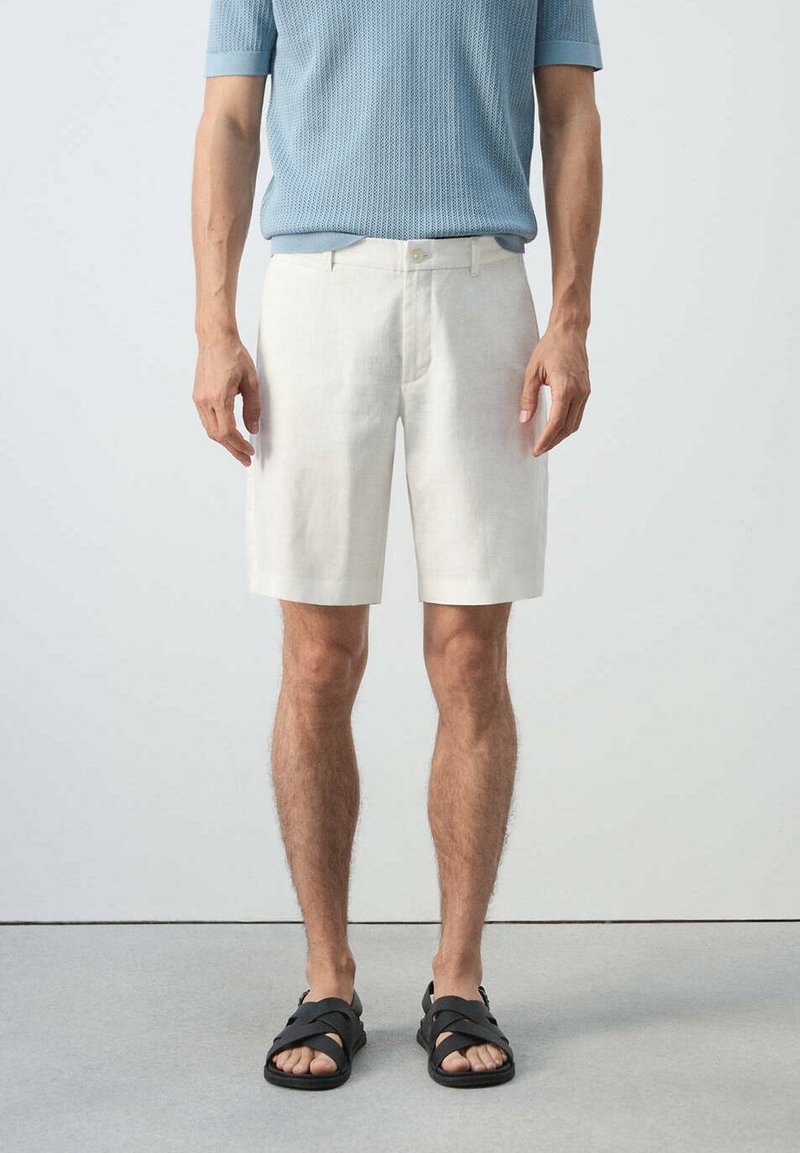 Homme portant une chemise en tricot bleu clair à manches courtes, un short blanc arrivant aux genoux et des sandales noires, debout devant un fond clair uni.