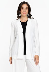 Blazer blanc à manches longues avec un col cranté et une fermeture à cinq boutons, associé à un top noir. Le tissu semble lisse avec une coupe décontractée.