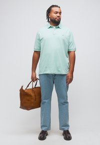 Άνδρας με πλεξούδες, που φοράει ανοιχτοπράσινο πόλο πουκάμισο και τζιν, κρατώντας μια μεγάλη καφέ δερμάτινη τσάντα ταξιδίου στο αριστερό του χέρι.