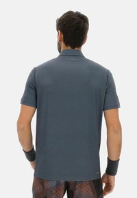 Man gekleed in een donkergrijze korte mouwen polo en gestreepte shorts, staande met zijn rug naar de kijker tegen een effen achtergrond.