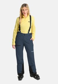Blauwe skibroek met verstelbare zwarte bretels, ritszakken en versterkte onderbenen, gecombineerd met een gele langemouwen shirt.