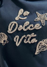 Mustmust kangas, millel on tekstuuritud beige helme tikandid, millel on ütlus "La Dolce Vita", koos dekoratiivsete meritähtede ja rannakarbi motiividega.