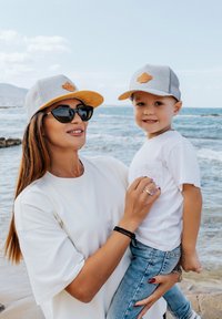 Két személy a tengerparton, egymással megegyező szürke sapkát viselnek, narancssárga részletekkel. A nő fehér pólót és napszemüveget hord, míg a gyermek fehér pólót és farmert visel.