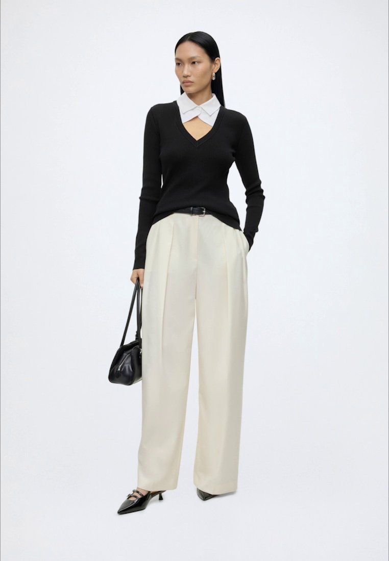 Femme debout portant un pull noir à col en V sur une chemise blanche à col, pantalon large beige, talons noirs, et tenant un sac à main noir.