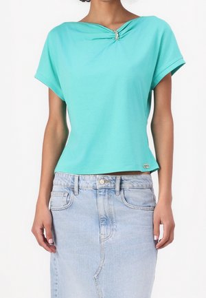 Femme portant un haut turquoise à manches courtes avec un petit ornement doré au niveau du col et une jupe en jean bleu clair.