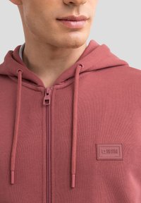 Roter Zip-Hoodie aus weichem Stoff, mit einem Logo-Patch auf der Vorderseite, geflochtenen Kordeln und strukturiertem Material.