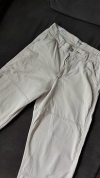 Calças de algodão bege com fecho de zíper, bolsos frontais e costuras planas. Textura suave com um ligeiro brilho, dispostas sobre um fundo escuro.