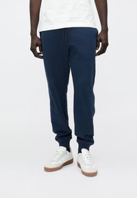 Man draagt marineblauwe joggers, witte sneakers met rubberen zolen en een wit shirt, staand tegen een effen achtergrond.