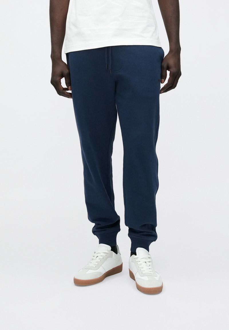 Man draagt marineblauwe joggers, witte sneakers met rubberen zolen en een wit shirt, staand tegen een effen achtergrond.