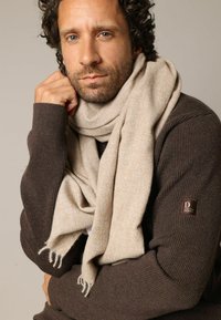 Écharpe en laine beige drapée autour du cou, portée avec un pull côtelé brun foncé. Détail notable de franges aux extrémités de l'écharpe.